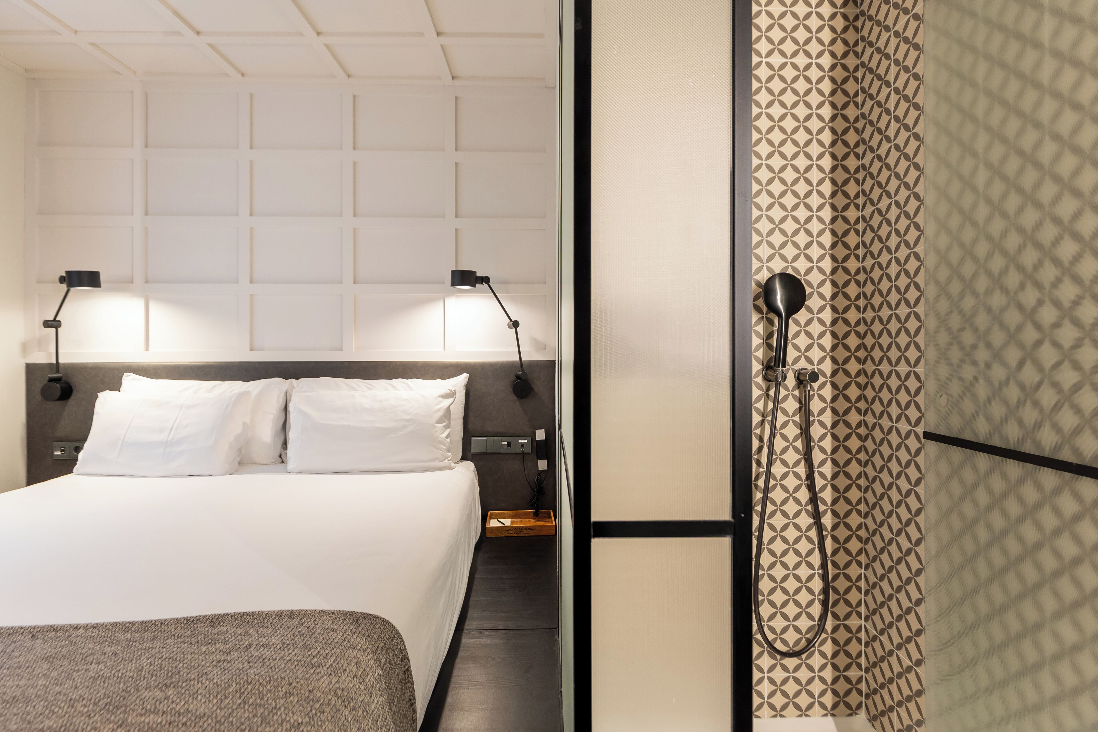 A hotel room with a neatly made bed, two bedside lamps, a nightstand, and a shower area with a patterned wall.