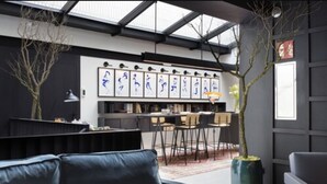 Un salon moderne avec un canapé, un coin repas avec des chaises, une grande fenêtre et un mur noir orné d’œuvres d’art encadrées.
