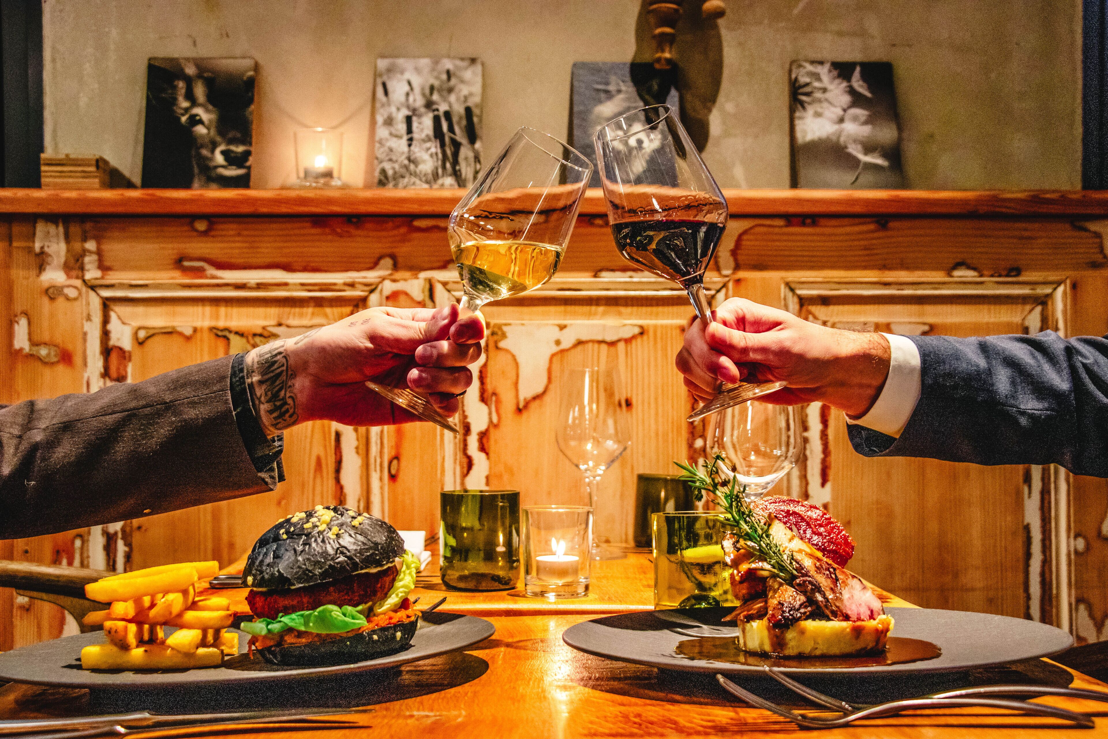Una mesa de madera con una hamburguesa, papas fritas y un filete con guarnición. Dos personas brindan con copas de vino.