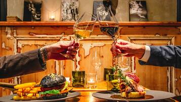 Una mesa de madera con una hamburguesa, papas fritas y un filete con guarnición. Dos personas brindan con copas de vino.