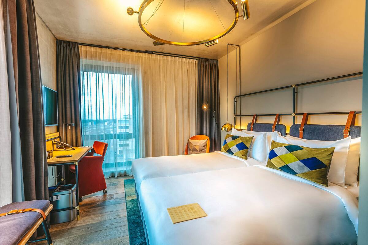 A hotel room with a large bed, a desk with a chair, a small table, and a view of the city through the window.