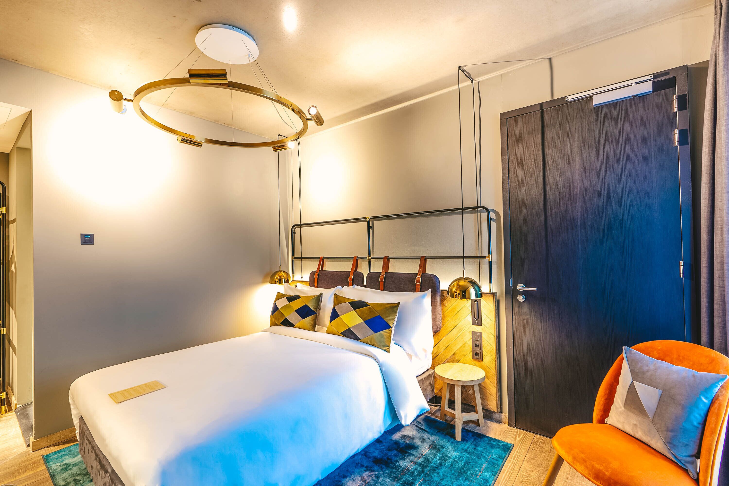 Een moderne hotelkamer met een groot bed, een klein krukje, een donkere deur en een bijzondere plafondlamp.