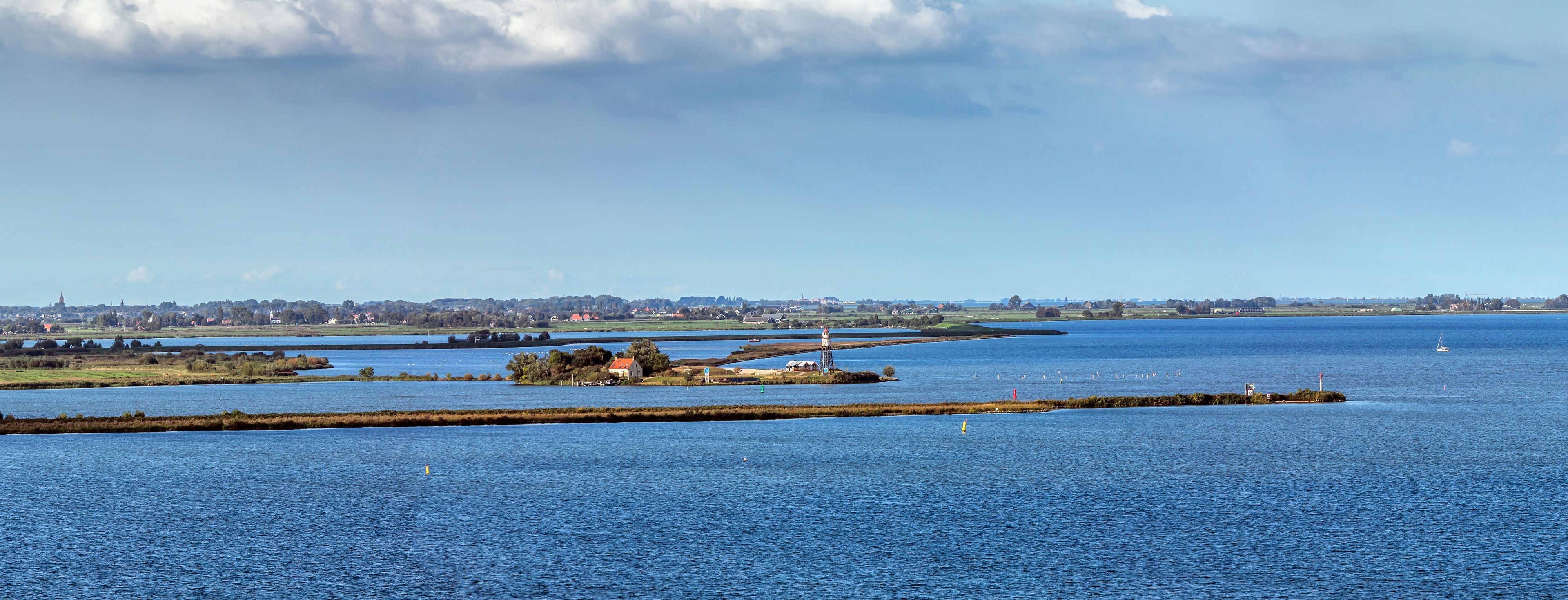 A coastal landscape with a lighthouse, calm water, and distant buildings.
