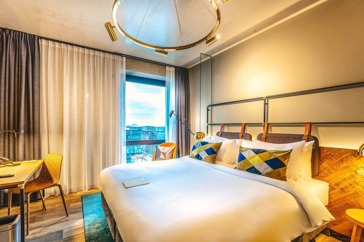 A hotel room with a large bed, a desk, a chair, and a view of the city through the window.