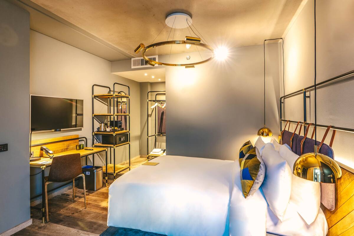 A modern hotel room with a large bed, a desk with a chair, a TV, and a wardrobe with hanging clothes.