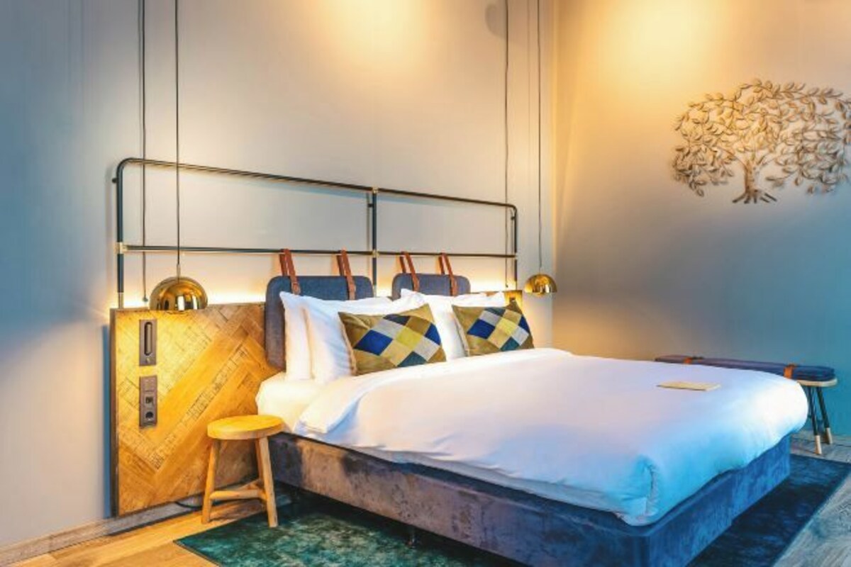 A modern bedroom with a bed, a wooden cabinet, a stool, and a wall-mounted light fixture.