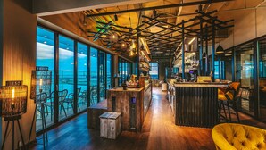 A modern bar area with a wooden bar counter, seating, and large windows offering a view of the water. - Four Elements Hotel Amsterdam (Amsterdam)