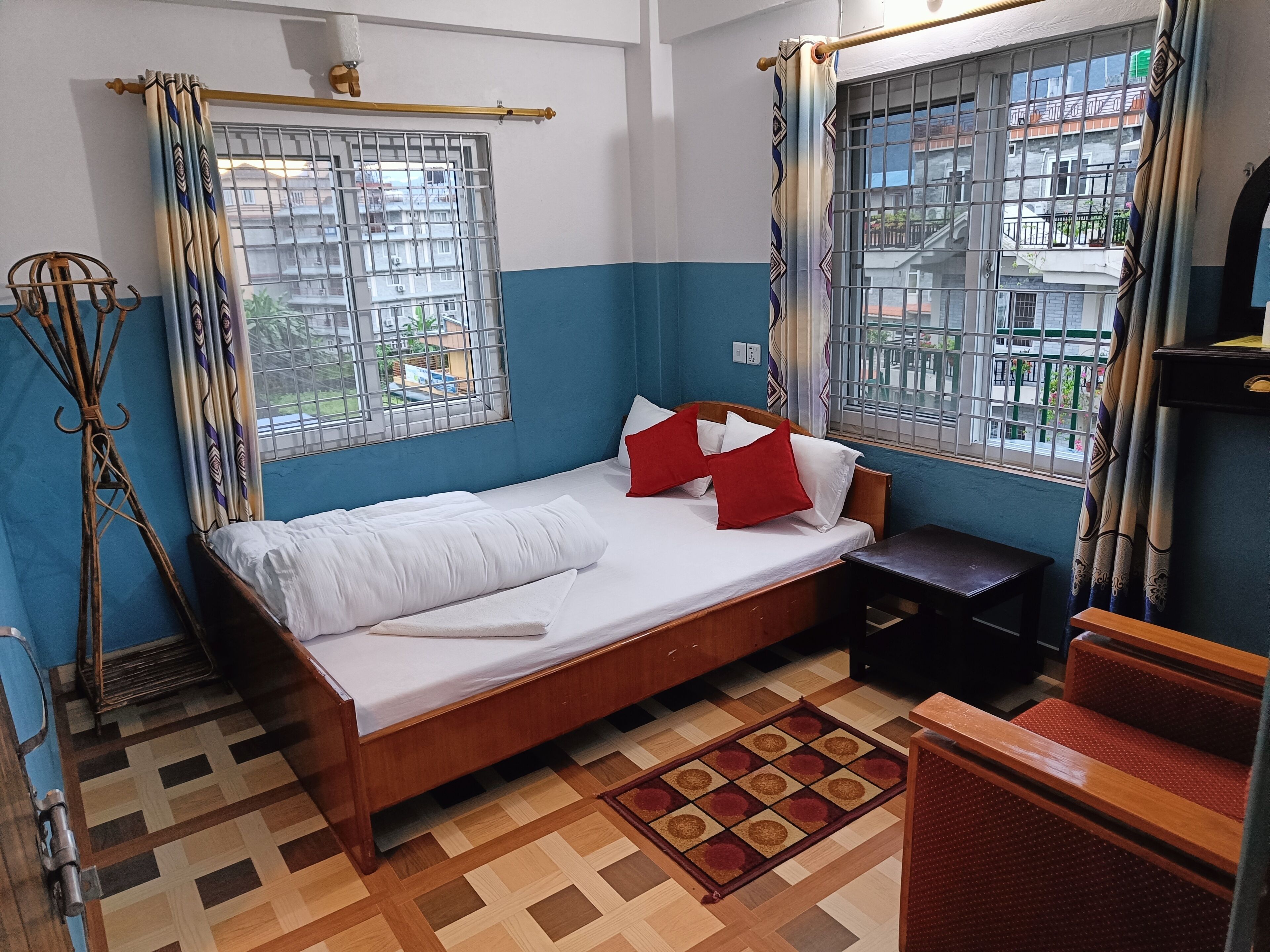 A bedroom with a wooden bed, a small bedside table, a patterned rug, and a view of the outside through barred windows.