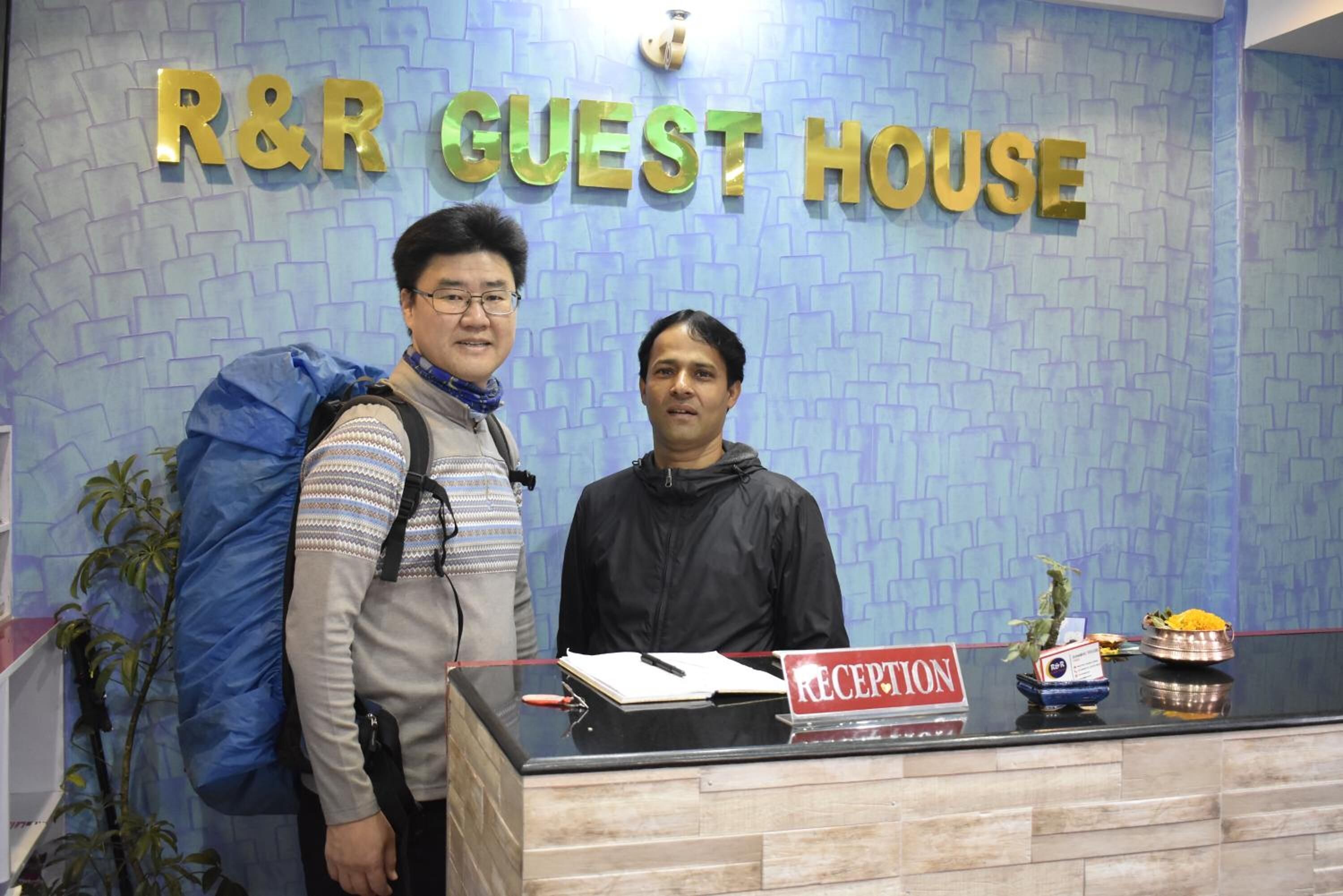 A reception desk with a sign reading "RECEPTION" and a backdrop with "R&R GUEST HOUSE" in gold letters.