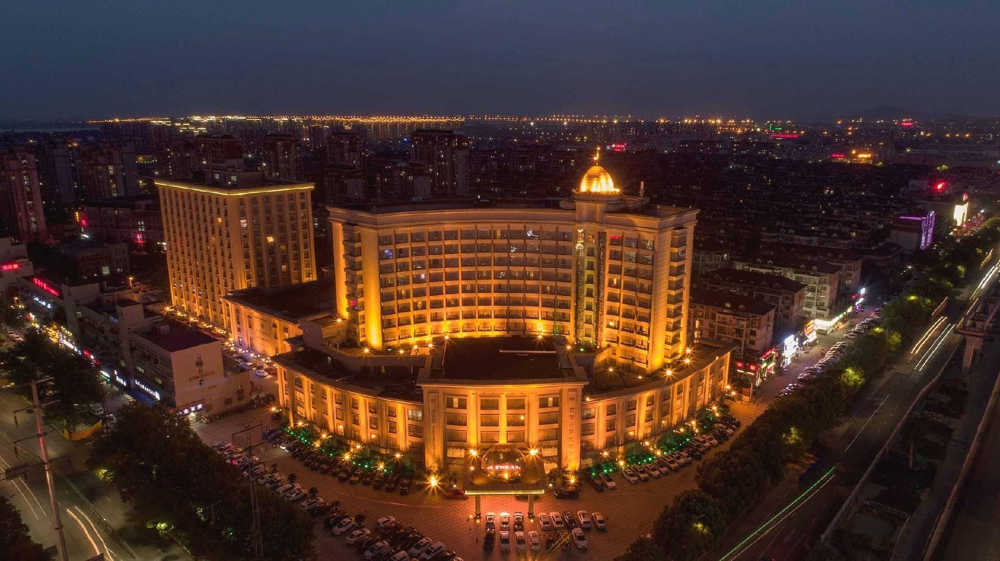 Un hotel de varios pisos con un patio central, rodeado por estacionamiento y carreteras.