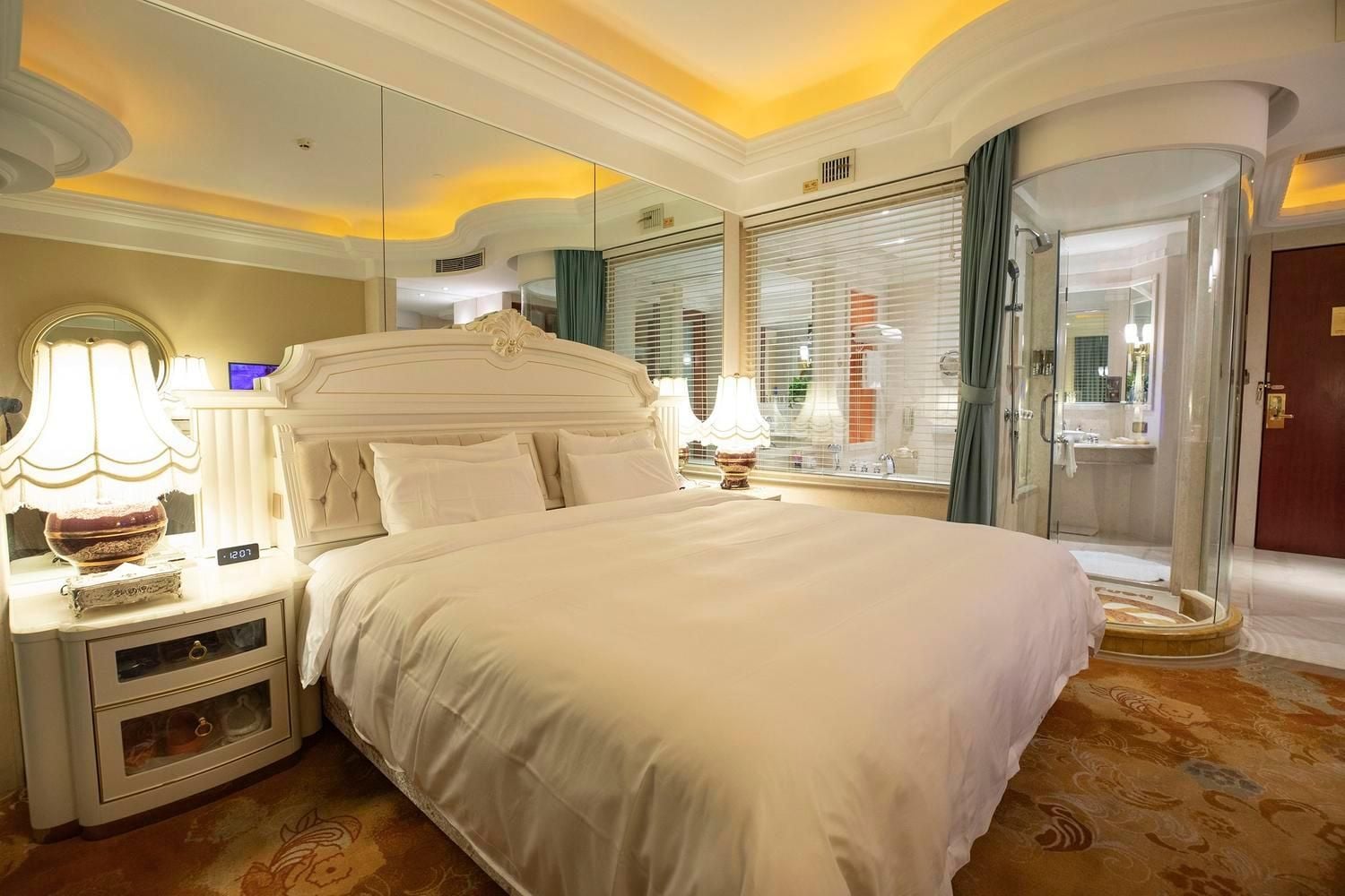 A hotel room with a large bed, a nightstand with a lamp, a mirror, and a glass-enclosed shower area.