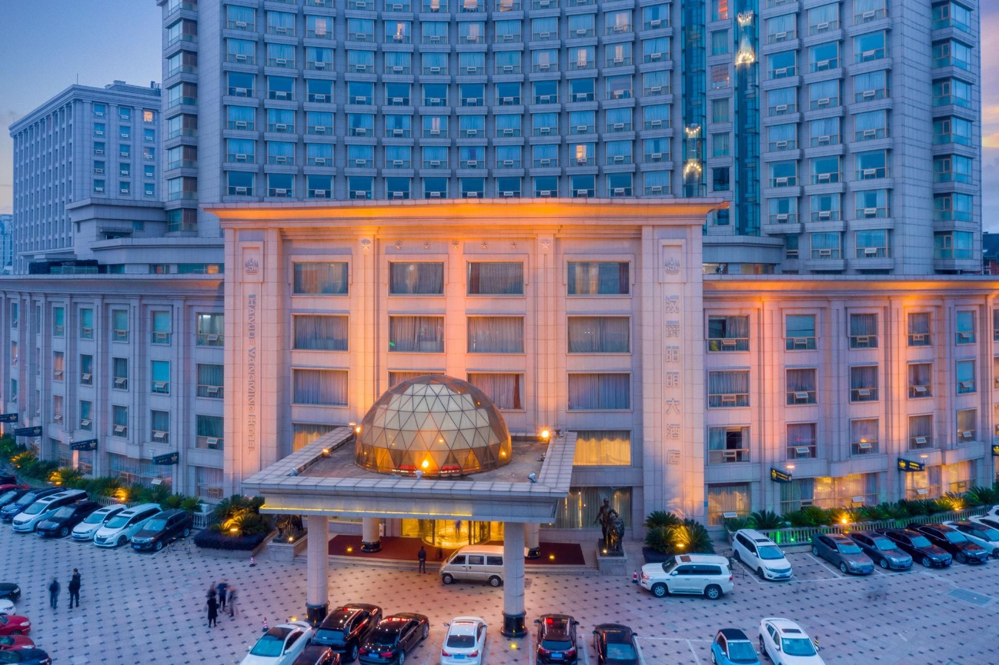 Un hotel con una estructura cúpula destacada en la entrada, varios pisos y un estacionamiento con varios autos.