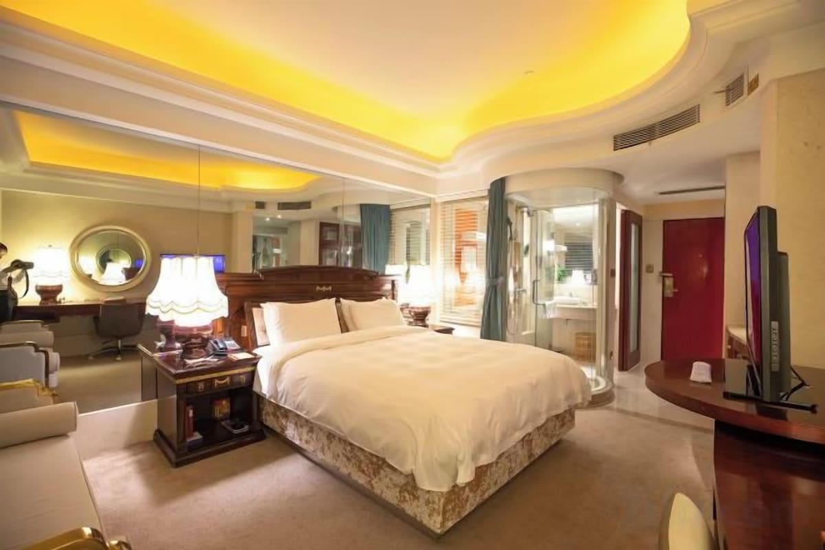 A hotel room with a large bed, bedside tables, a desk, a chair, a mirror, and a TV.
