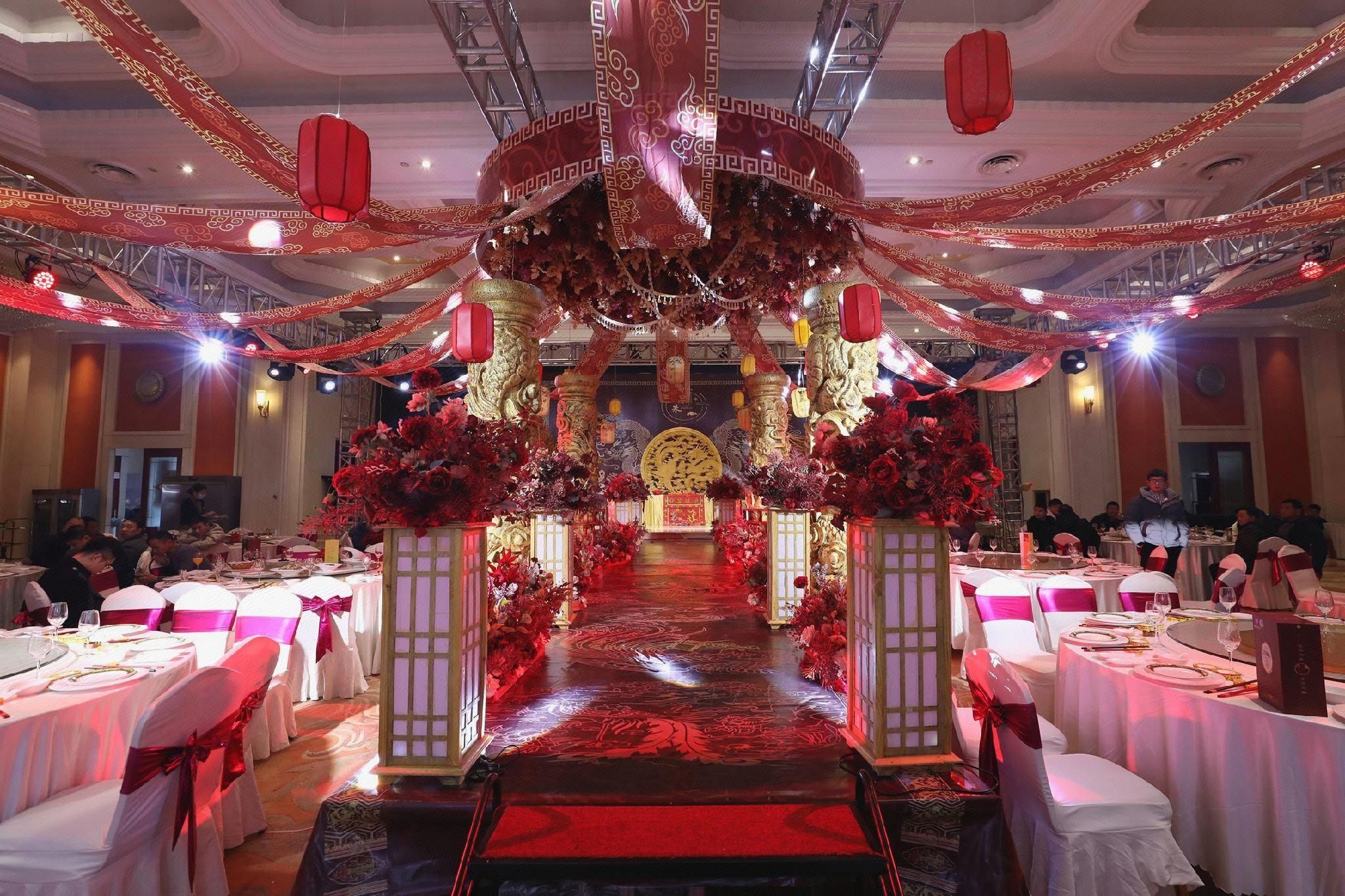 A banquet hall with tables set for a formal event, a central aisle leading to a decorated stage, and red lanterns hanging from the ceiling.