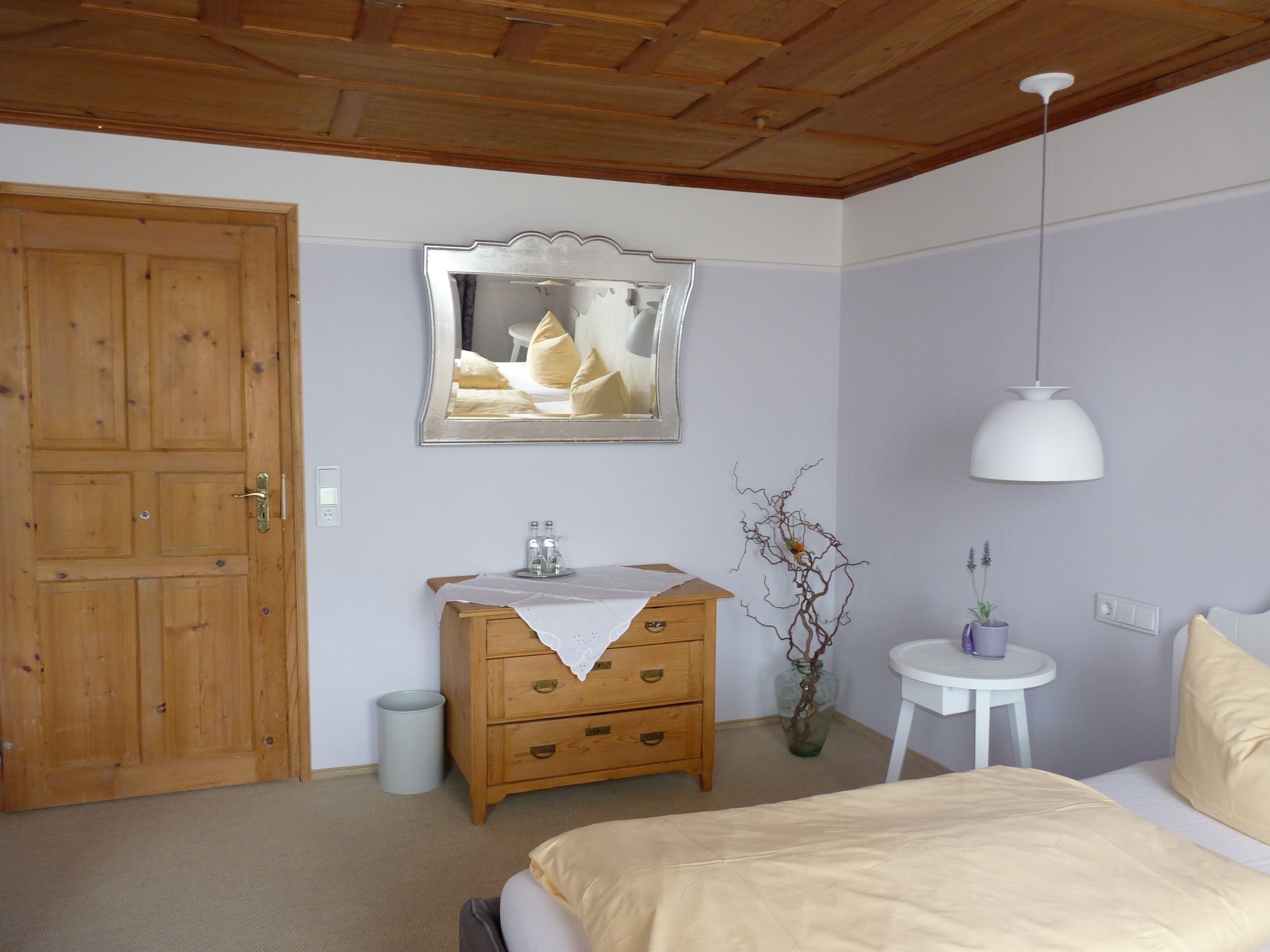Una camera da letto con una porta in legno, una cassettiera con specchio, un comodino, una lampada a sospensione e un letto con coperta beige.