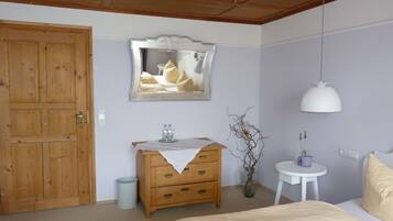 Una camera da letto con una porta in legno, una cassettiera con specchio, un comodino, una lampada a sospensione e un letto con coperta beige.