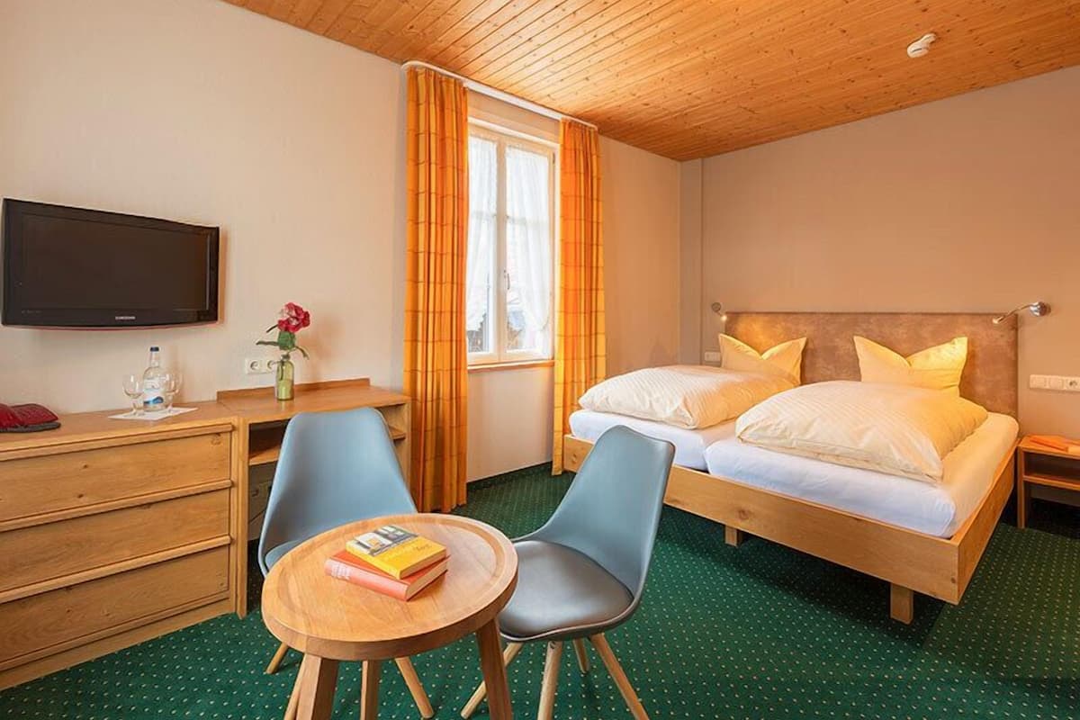 Une chambre d’hôtel avec un plafond en bois, un lit avec une literie blanche, un bureau avec une chaise, une télévision et une fenêtre avec des rideaux.