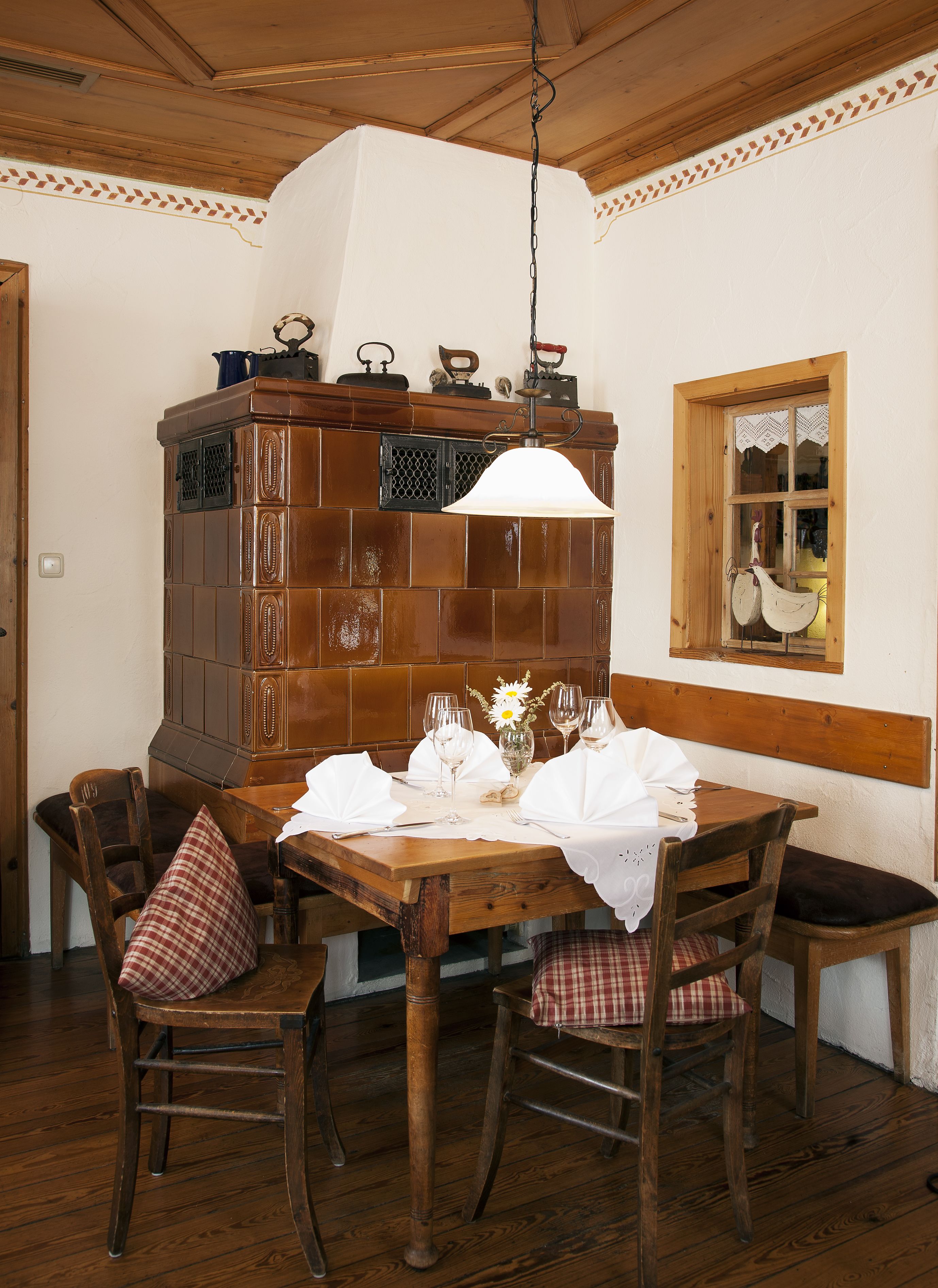 A dining area with a wooden table set for four, surrounded by wooden chairs with checkered cushions. A hanging light fixture is above the table, and a wooden cabinet with a tiled backsplash is to the left.