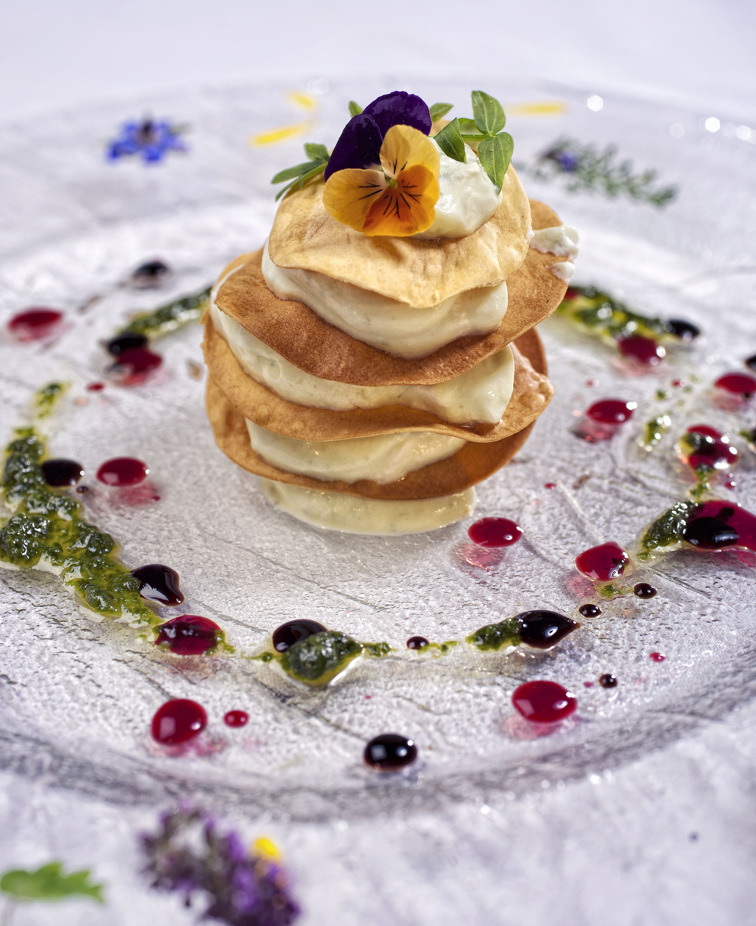 Uma pilha de panquecas com uma flor decorativa por cima, servida em um prato com açúcar de confeiteiro e molho de frutas vermelhas.