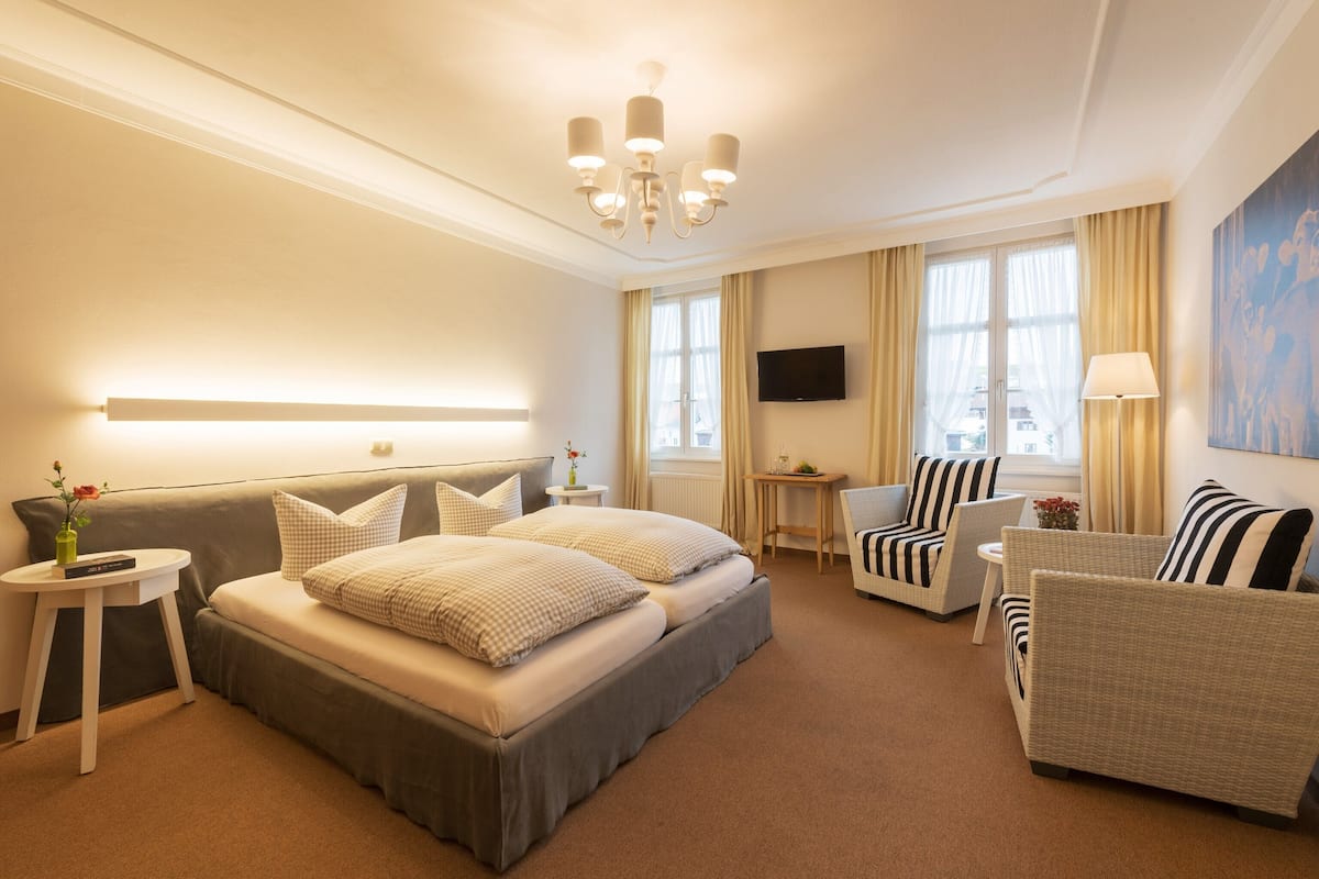 Une chambre d’hôtel avec un grand lit, deux fauteuils, une petite table, un téléviseur fixé au mur et une fenêtre avec des rideaux.