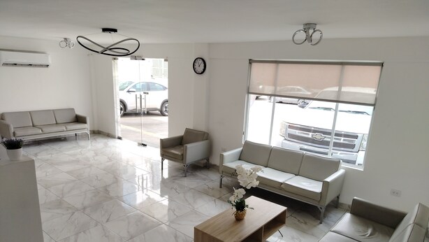 Un salón moderno y bien iluminado con suelo de mármol, un sofá color beige, una mesa de centro de madera y vistas a coches aparcados a través de amplios ventanales.