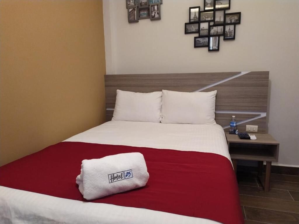 A hotel room with a neatly made bed, a red blanket, a white towel with a hotel logo, a nightstand with a water bottle, and a wall with framed pictures.
