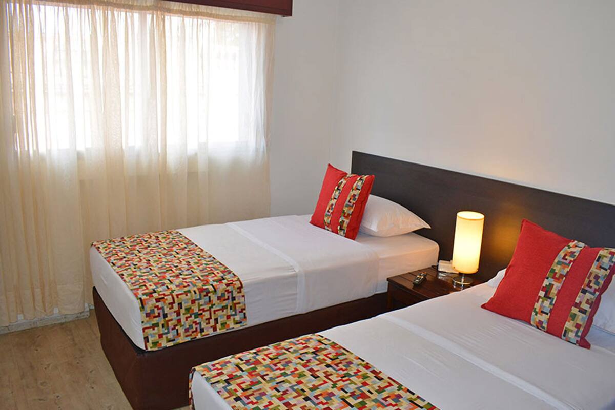 Habitación de hotel con dos camas, cada una con una colcha estampada y almohadas rojas. Hay una mesita de noche con una lámpara entre las camas.