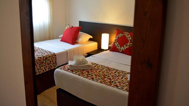 A hotel room with two beds, each with a patterned bedspread and red pillows. There is a nightstand with a lamp between the beds. - Hotel Express (Maldonado)