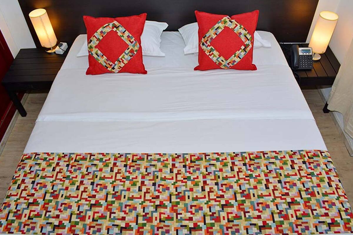 Una cama bien tendida con sábanas blancas y dos almohadas rojas con un diseño geométrico. Hay dos mesitas de noche con lámparas, un teléfono y una pequeña mesa con una lámpara.
