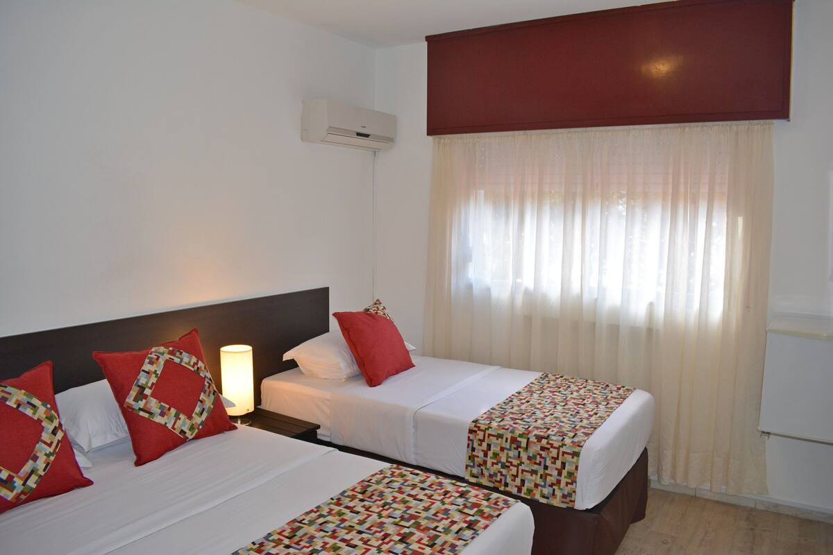 Habitación de hotel con dos camas, cada una con una colcha estampada y almohadas rojas. Se observa una mesita de noche con una lámpara, una ventana con cortinas traslúcidas y un equipo de aire acondicionado en la pared.