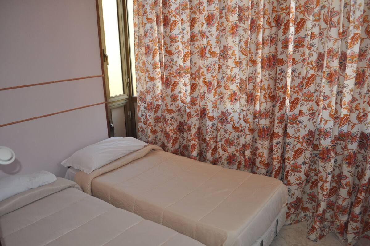 A room with two beds, a window, and floral curtains.