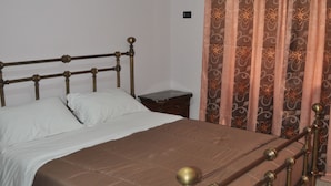 A bed with a brown headboard and footboard, a white mattress, and a brown bedspread. There is a nightstand with a lamp and a small table beside the bed. A patterned curtain is partially drawn, and an air conditioning unit is mounted on the wall above the bed. - Naples Experience B&B (Naples)