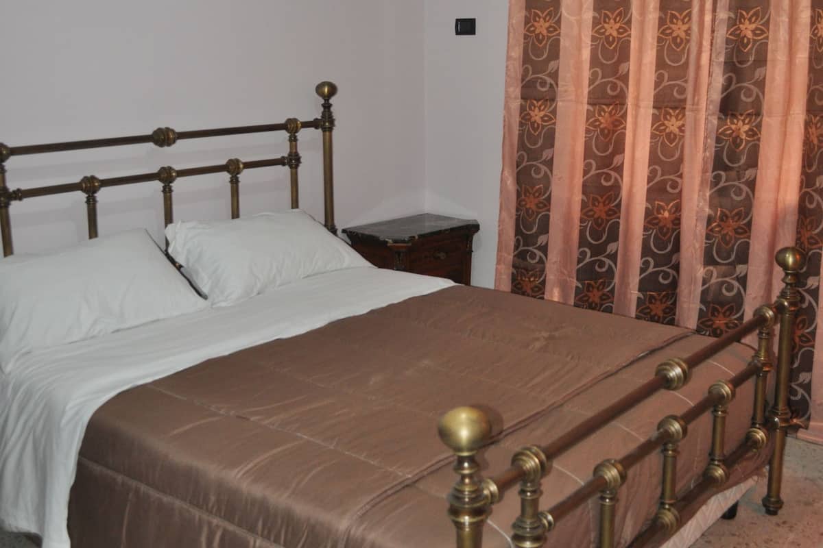 A bed with a brown headboard and footboard, a white mattress, and a brown bedspread. There is a nightstand with a lamp and a small table beside the bed. A patterned curtain is partially drawn, and an air conditioning unit is mounted on the wall above the bed.