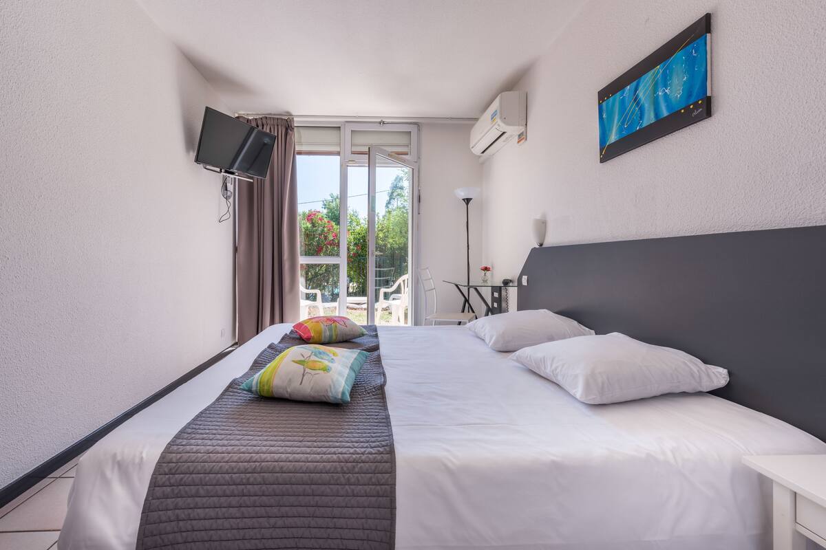 Une chambre d’hôtel avec un lit, un téléviseur fixé au mur, un ventilateur de plafond et une fenêtre avec des rideaux.