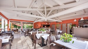 Un restaurant spacieux avec de hauts plafonds, des poutres blanches et un mélange d’espaces de restauration intérieurs et extérieurs. Les tables sont dressées avec des nappes et des chaises blanches, et des plantes en pot y sont disposées.