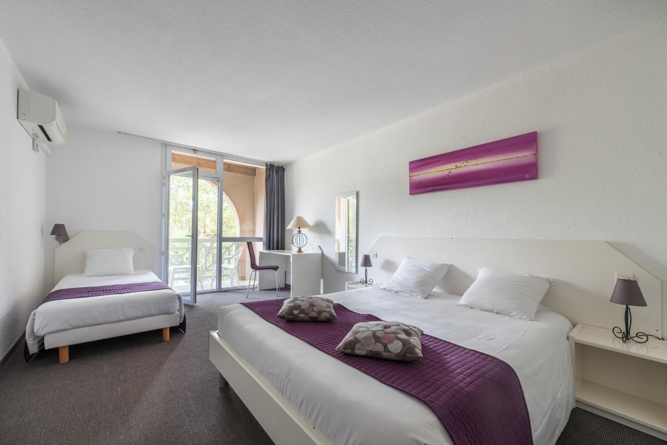 Ein Hotelzimmer mit einem großen Bett, einem kleineren Bett, einem Schreibtisch und einem Sessel. Es gibt ein Fenster mit Blick auf Grünflächen.