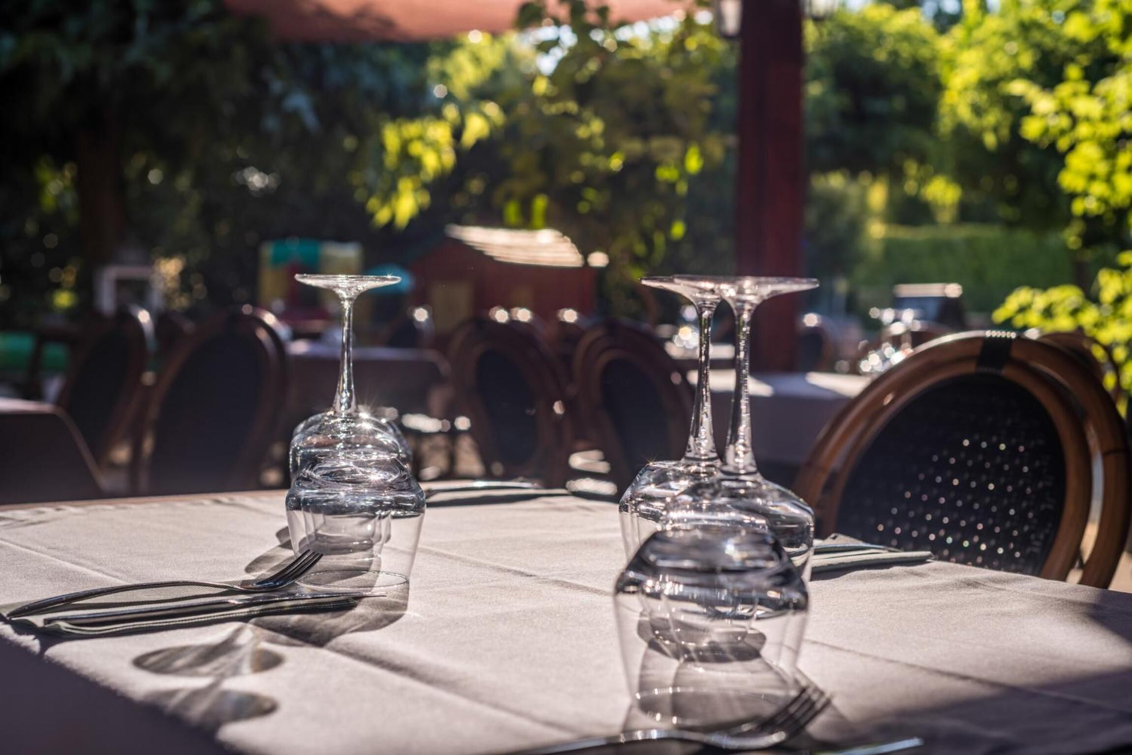 A table set with clear glassware and cutlery, with a view of outdoor seating and greenery in the background.