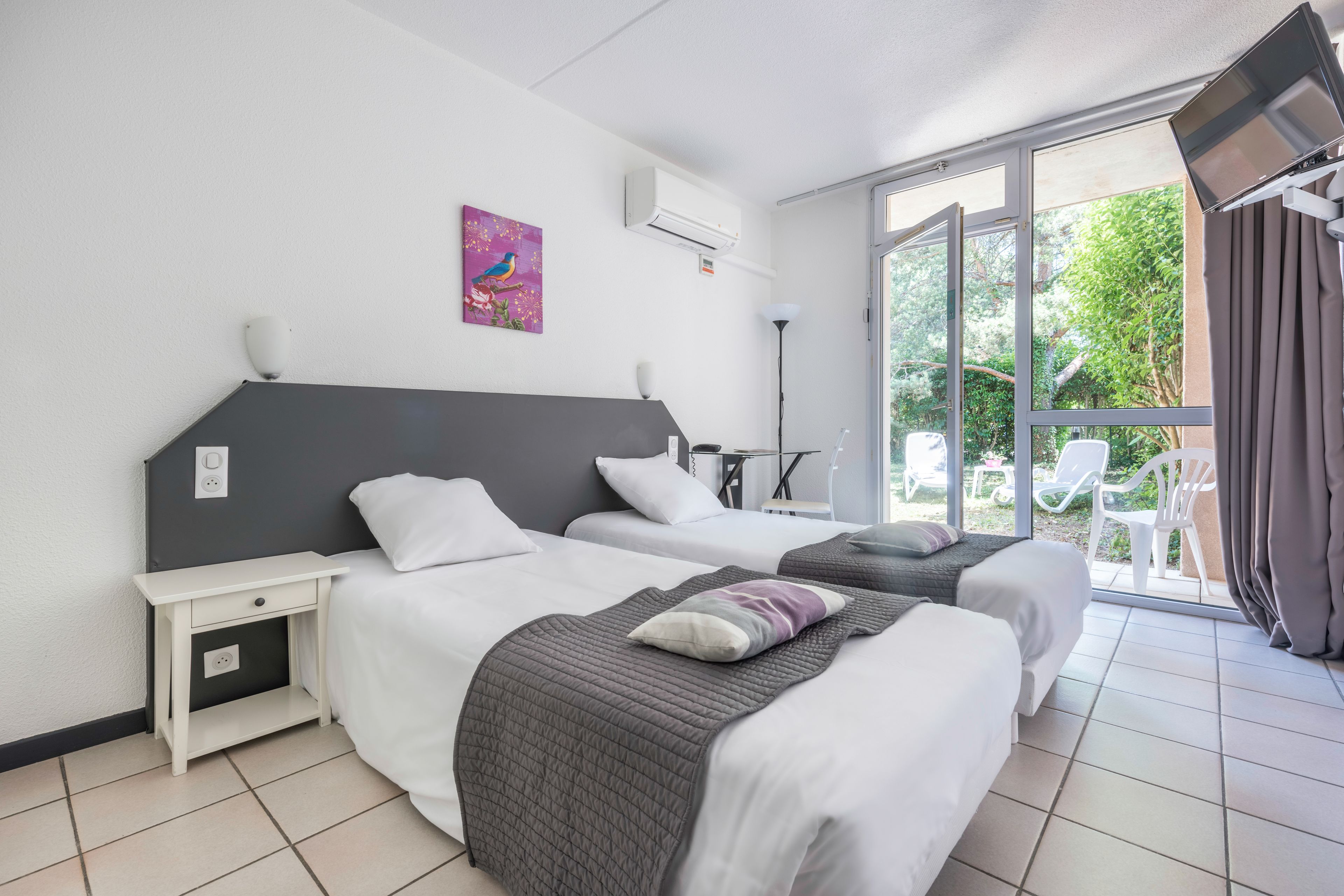 A hotel room with two beds, a small bedside table, a wall-mounted air conditioner, a window with a view of greenery, and a ceiling-mounted TV.