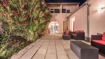 Un patio exterior con un camino que lleva a la entrada de un edificio, amueblado con asientos negros y rojos.