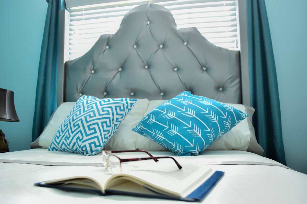 A bed with a tufted headboard, two patterned pillows, and an open book on the bed.