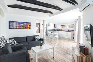 Un salon moderne avec un canapé, une table basse sur laquelle se trouvent une bouteille et des verres, un téléviseur, et une cuisine en arrière-plan.