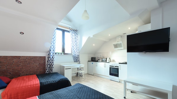 Un appartement compact doté d’une kitchenette, d’un lit avec une literie rouge et bleue, d’un téléviseur fixé au mur et d’une fenêtre ornée de rideaux à pois.