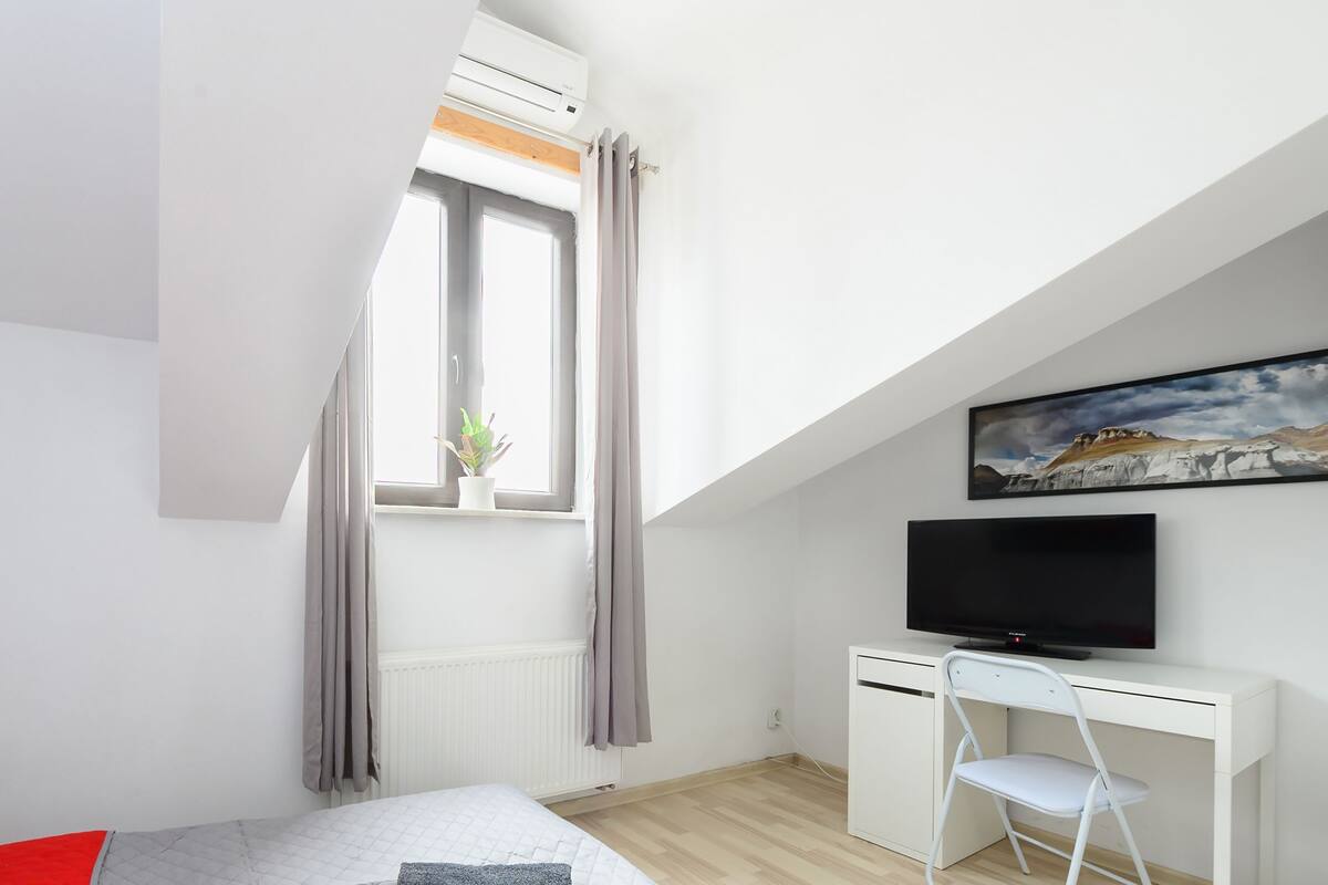 Un dormitorio moderno con cama, escritorio, silla, televisor y ventana con cortinas.