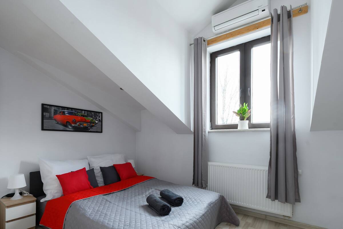 Un dormitorio con cama, mesita de noche, lámpara, ventana con cortinas, radiador y aire acondicionado instalado en la pared.