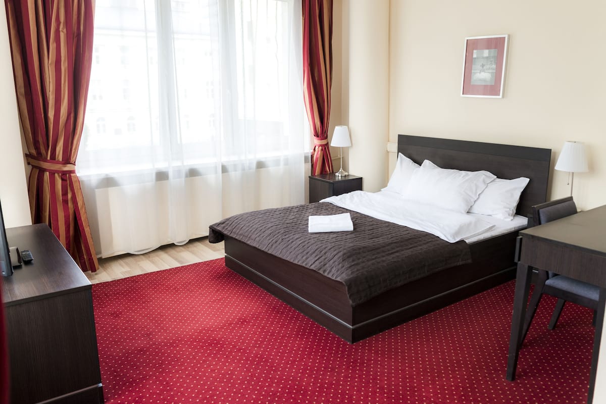 Habitación de hotel con una cama grande, un escritorio y una silla. Hay dos mesitas de noche con lámparas. La habitación tiene una alfombra roja y cortinas.