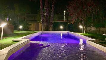 Una piscina iluminada con luces moradas durante la noche, rodeada de árboles y una valla bien iluminada.