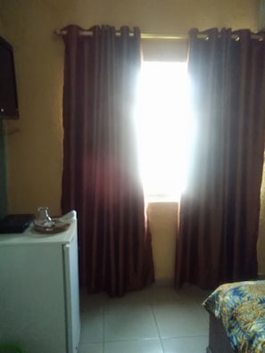 A room with a bed, a small table with a lamp, and a window with curtains. - Kingsby homes and hotel (Accra)