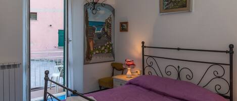 A bedroom with a bed, a nightstand, a chair, a table, a painting, and a view through an open door.