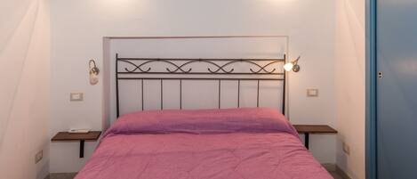 Una cama bien hecha con una colcha rosa y almohadas blancas, dos mesitas de noche con lámparas, y una puerta azul.