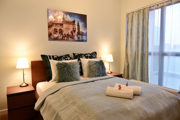 Una cama bien hecha con un edredón acolchado, dos almohadas mullidas y un par de toallas dobladas. Hay una mesita de noche con una lámpara encima, y un cuadro de un edificio histórico en la pared.